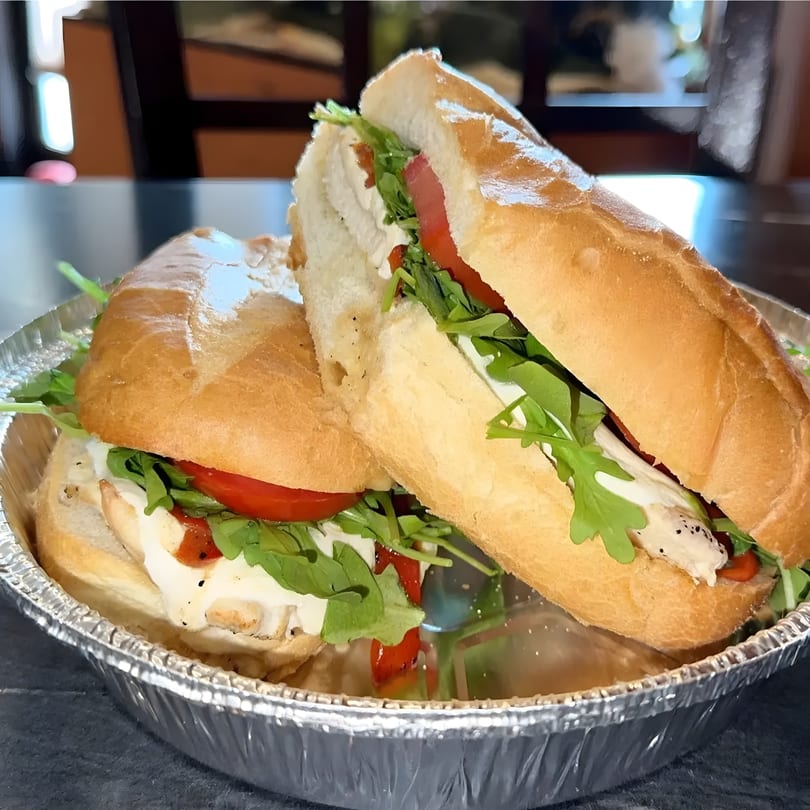 Two halves of a baguette sandwich with chicken, mozzarella, arugula, and roasted red peppers in a foil container.