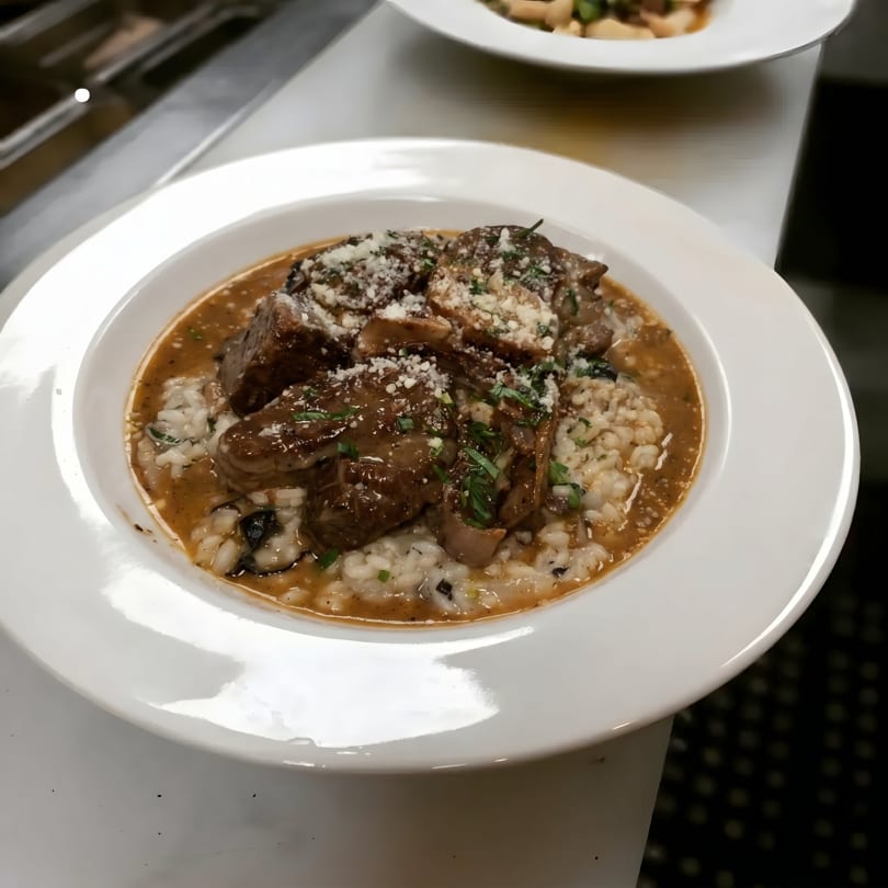Braised short ribs with creamy risotto, garnished with grated cheese and fresh herbs in a white bowl.
