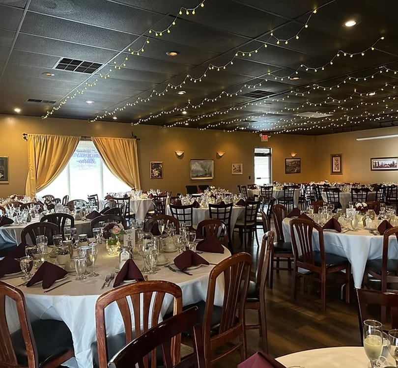 A restaurant dining room with round tables set with white tablecloths, maroon napkins, and string lights.
